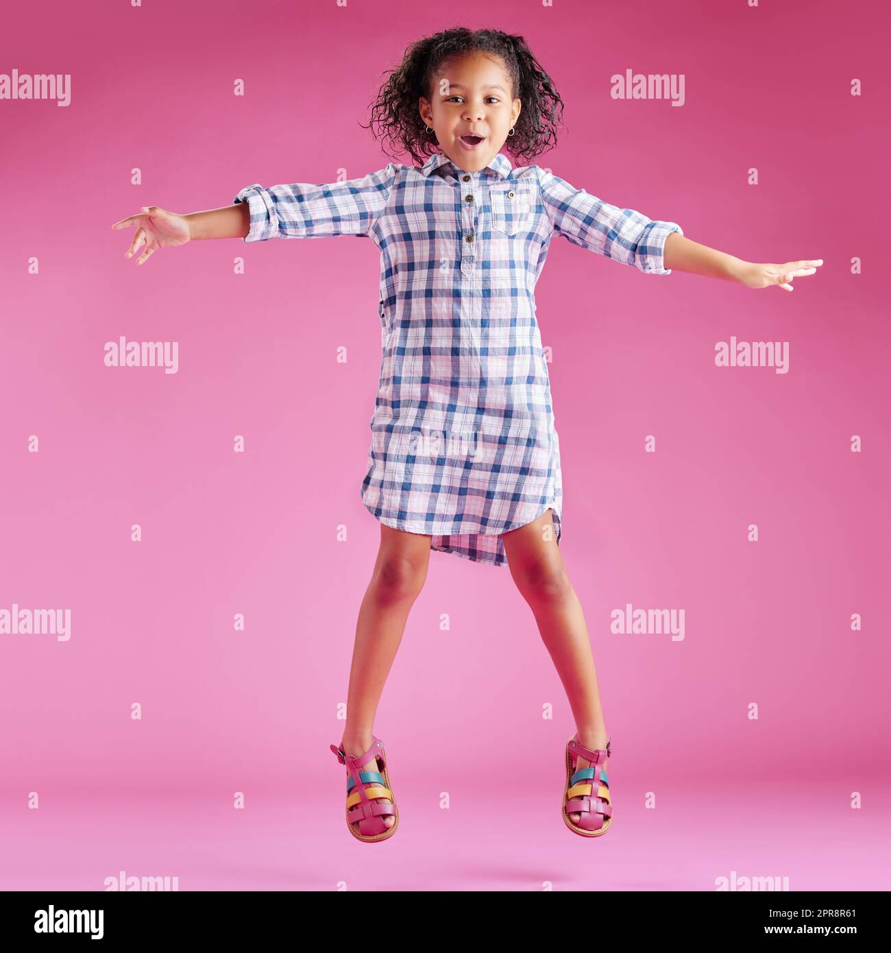 Ein hübsches, gemischtes Rennmädchen mit lockigem Haar feiert und tanzt vor einem pinkfarbenen Hintergrund in einem Studio. Afrikanisches Kind springt und fühlt sich frei Stockfoto