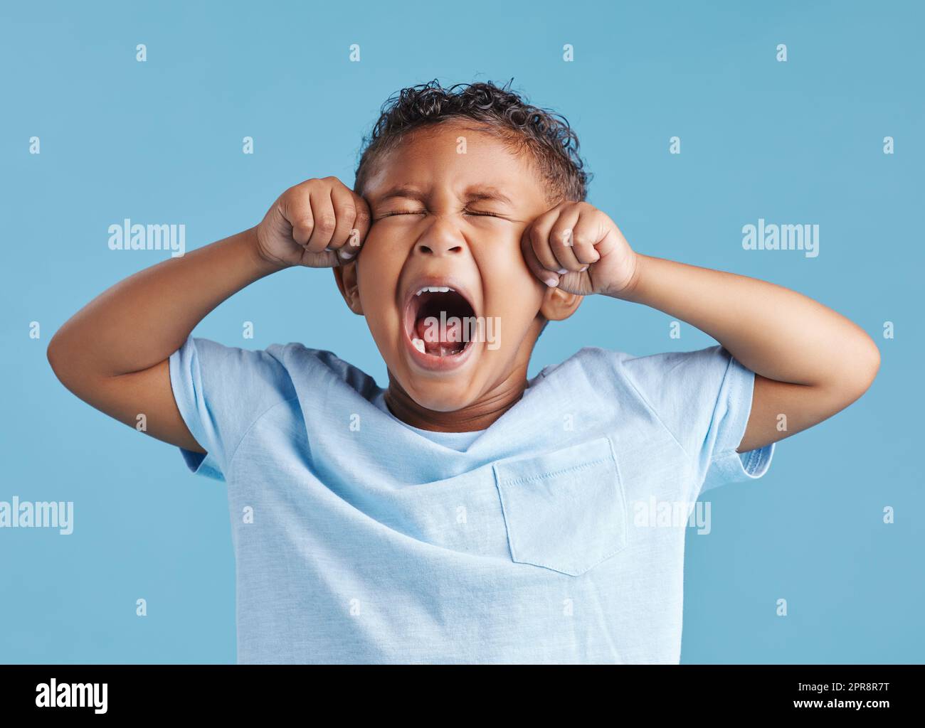 Unglücklicher kleiner lateinamerikanischer Junge, der aufgebracht aussah und weinte, während er seine Augen vor einem blauen Studiohintergrund reibt. Unglücklicher Vorschulkinder, der sich die Augen ausweint Stockfoto