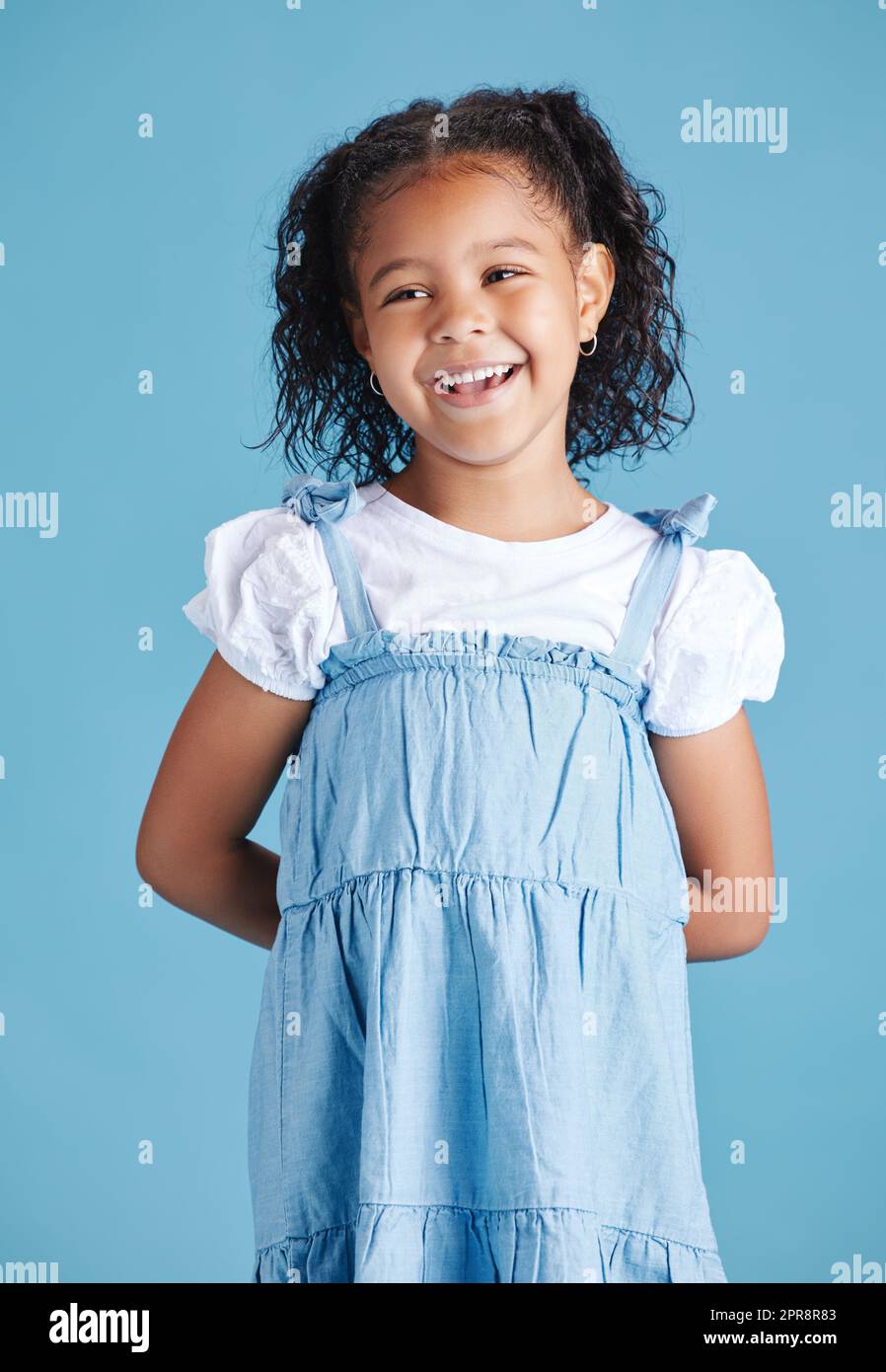 Fröhliches, lächelndes kleines Mädchen, das mit den Händen hinter dem Rücken vor blauem Studiohintergrund steht. Fröhliches Mischling-Kind in legerem Jeanskleid und weißem T-Shirt Stockfoto