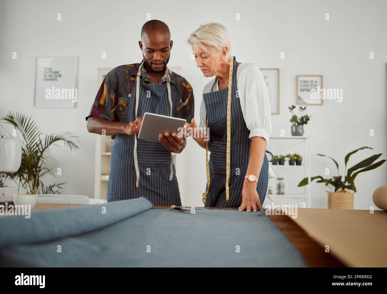 Zwei Bekleidungsdesigner, die ein digitales Tablet verwenden, während sie mit Material arbeiten. Junger afroamerikanischer Schneider, der mit einer Kaukasierin zusammenarbeitet und ein digitales Tablet in der Hand hält Stockfoto