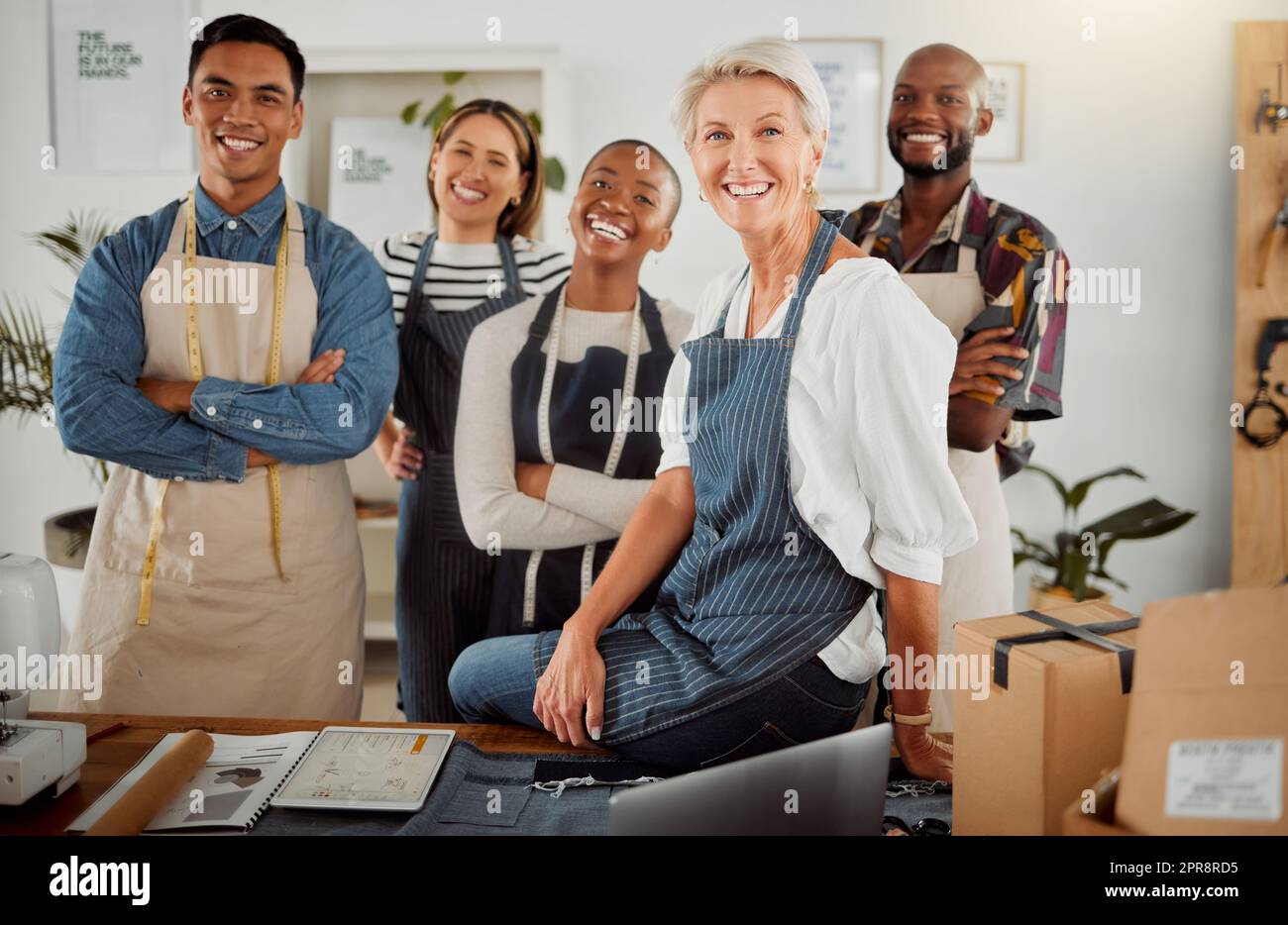 Eine Gruppe von fünf fröhlichen, vielfältigen Bekleidungsdesignern, die in einem Geschäft auf der Arbeit mit gekreuzten Armen stehen. Fröhliche Schneider, die lächeln und lachen, während sie zusammen in einer Boutique stehen. Reife, weiße Chefin, die bei ihren Angestellten steht Stockfoto