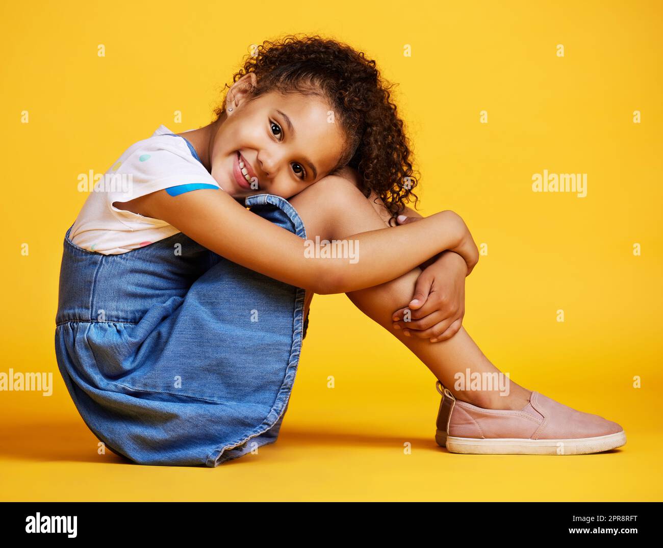 Studio-Porträt gemischtes Rassenmädchen, das allein vor gelbem Hintergrund sitzt. Süßes hispanisches Kind, das drinnen posiert. Fröhliches und süßes Kind, das lächelt und sorgenfrei aussieht in lässiger Kleidung Stockfoto