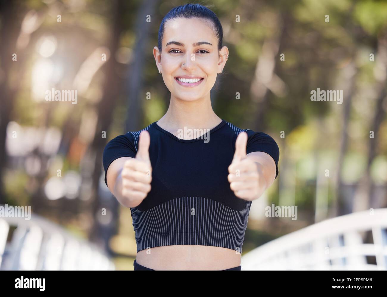 Glückliche Sportlerin, die beim Laufen oder Joggen im Freien mit beiden Händen die Daumen nach oben zeigt. Fit, junge Frau, die beim Workout im Park lächelt und zufrieden aussieht Stockfoto