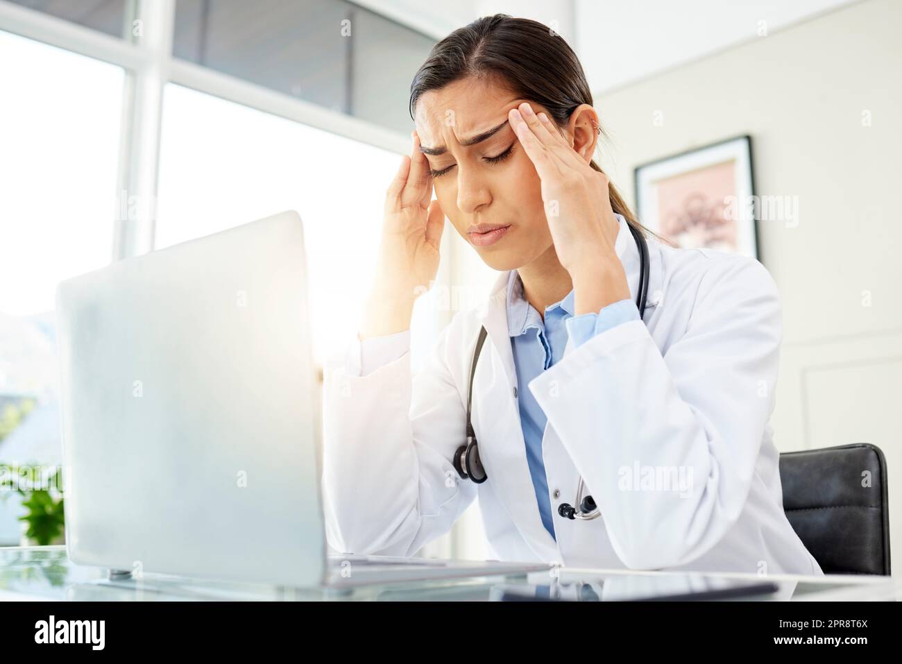 Nahaufnahme einer jungen gemischten Ärztin, die besorgt aussah und Kopfschmerzen hatte, während sie an einem Computer in ihrem Büro arbeitete. Hispanische Frau mit Kopfschmerzen und Stress im Krankenhaus Stockfoto