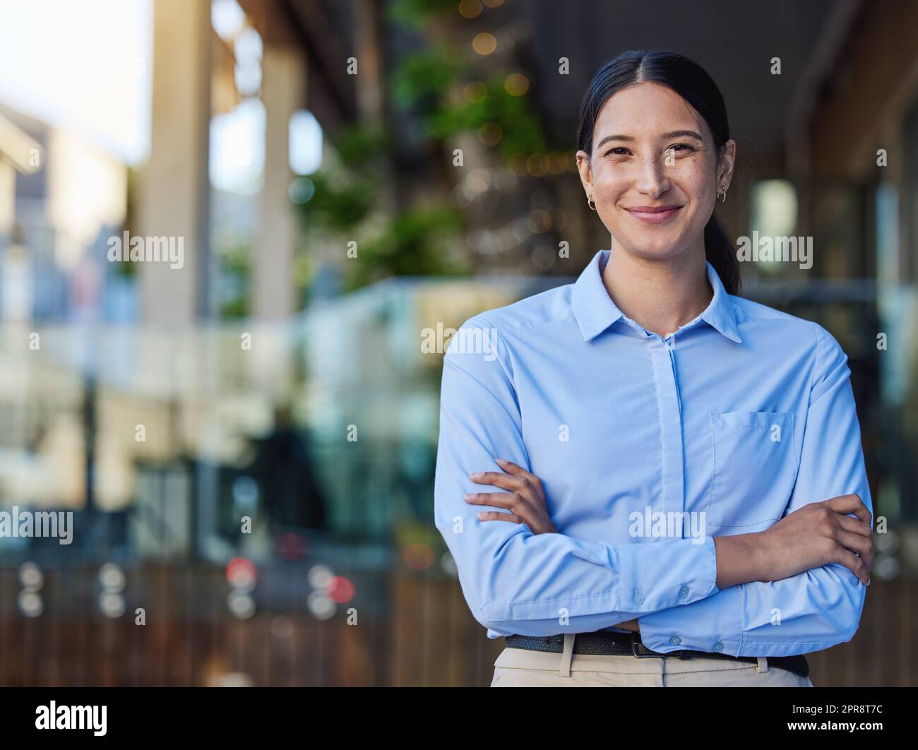 Porträt einer fröhlichen gemischten Rasse, lächelnde Geschäftsfrau mit nach außen gefalteten Armen. Junge hispanische Geschäftsführerin steht draußen vor verschwommenem Werbespot Stockfoto