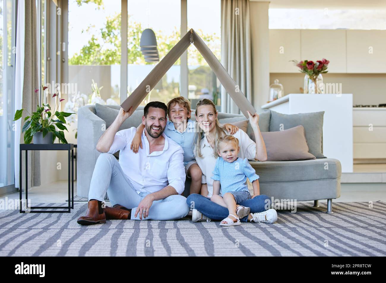 Waren versichert. Das ganze Porträt einer liebevollen vierköpfigen Familie, die ein Pappdach über den Kopf hält, während sie zu Hause im Wohnzimmer sitzen. Stockfoto