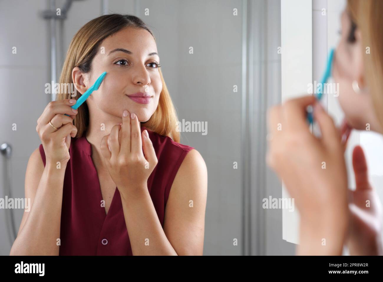 Entfernung von Gesichtshaaren. Brasilianisches Mädchen, das sich zu Hause mit einem Rasiermesser ins Gesicht rasiert. Schöne junge Frau mit Rasiermesser auf Bad. Stockfoto