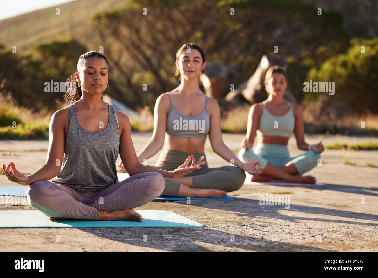 Lange Yoga-Frauen meditieren mit gekreuzten Beinen für Outdoor-Übungen in abgelegener Natur. Vielfältige Gruppe von aufmerksamen, aktiven Freunden, die psychische Gesundheit verbinden und ausbalancieren. Junge, fokussierte, ruhige Zen-Leute Stockfoto