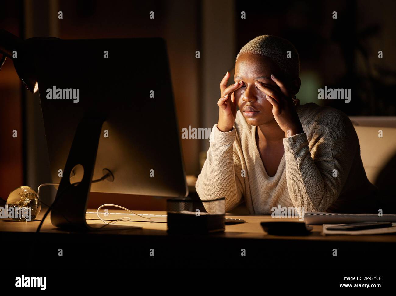 Meine Augen wollen einfach nur geschlossen bleiben. Eine junge Geschäftsfrau, die gestresst schaut, während sie nachts an einem Computer in einem Büro arbeitet. Stockfoto