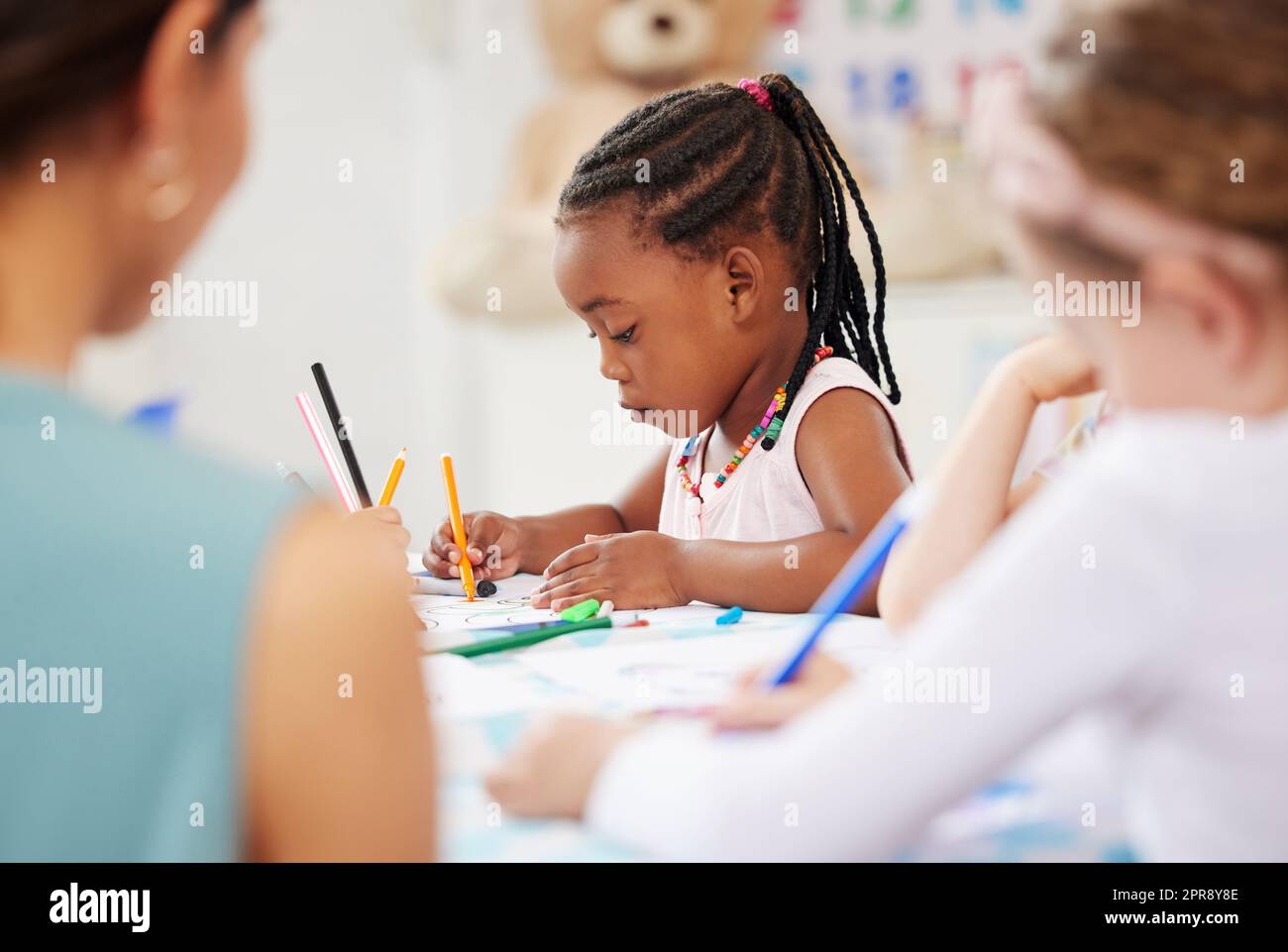 Ein afroamerikanisches Mädchen, das mit seinen Mitschülern an einem Tisch sitzt und im Vorschulalter oder Kindergarten malt. Eine junge Frau, die in ihrer Schule mit bunten Bleistiften Bilder in einem Klassenzimmer malt Stockfoto