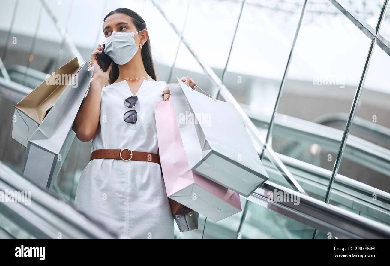 Eine junge Mischrasse Frau trägt eine medizinische Gesichtsmaske und spricht auf einem Handy während einer Rolltreppe nach einem Einkaufsbummel. Modischer hispanic mit Einkaufstaschen nach Kauf in einem Einkaufszentrum während der Covid-19-Pandemie Stockfoto