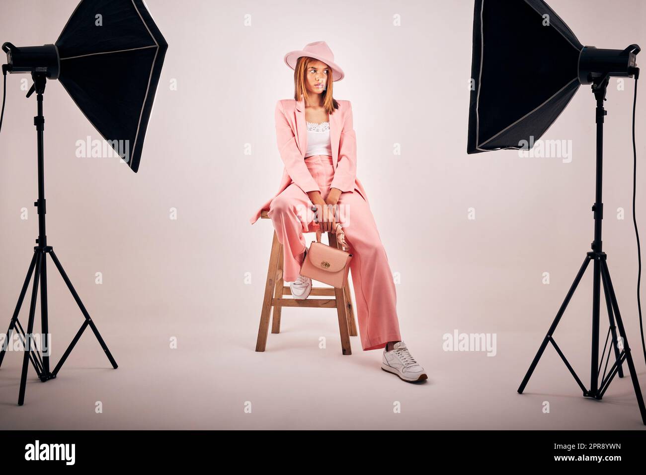 Porträt einer jungen Mischrasse, die in trendiger modischer Kleidung posiert und auf einem Stuhl mit grauem Studiohintergrund chillt. Hispanische Frau in der neuesten Modesammlung mit coolem Stil und Hut Stockfoto