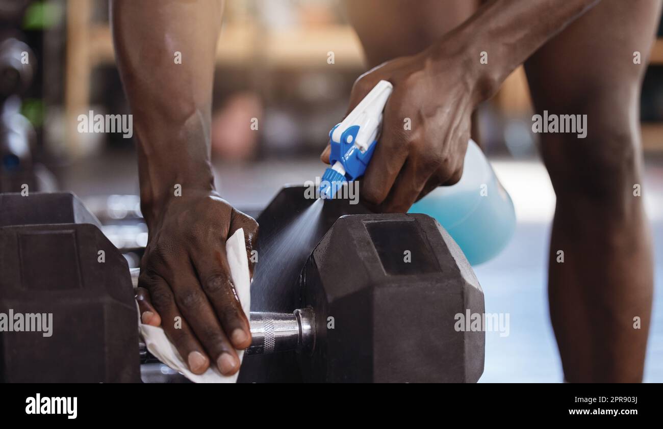 Nahaufnahme eines unbekannten afroamerikanischen Athleten mit einer Sprühflasche zur Reinigung von Kurzhanteln im Fitnessstudio. Aktive Black-man-Reinigungsgeräte im Hygienealltag zum Schutz und zur Vermeidung von Covid durch körperliche Betätigung Stockfoto