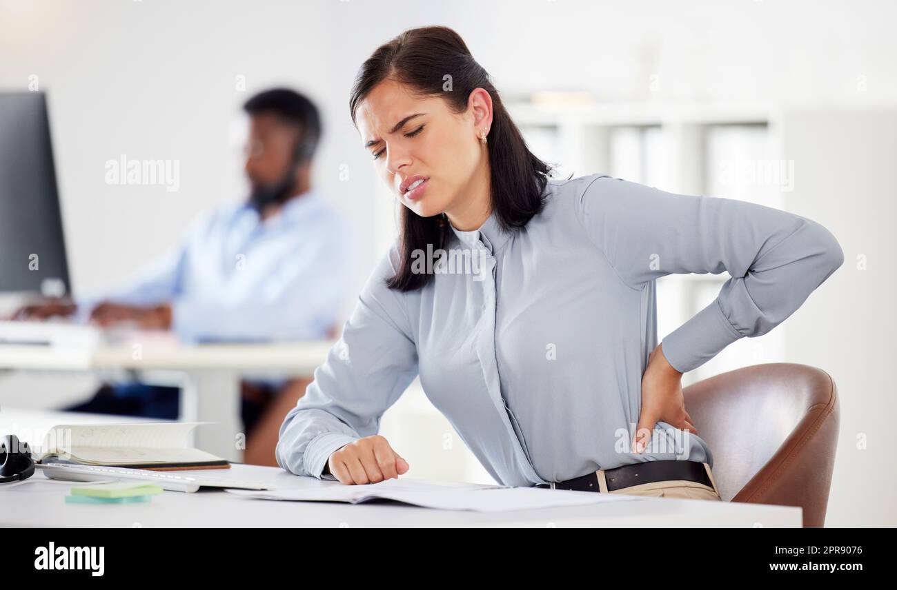Eine junge, gestresste, weiße Geschäftsfrau, die unter Rückenschmerzen leidet, in einem Büro. Weibliche Mitarbeiterin, die sich angespannt fühlt, sich unwohl fühlt und sich mit einer schlechten Sitzhaltung und langen Arbeitszeiten am Schreibtisch verletzt Stockfoto