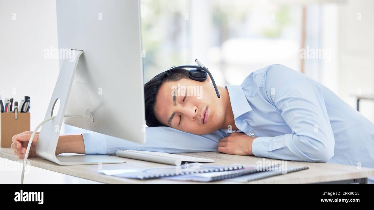 Nahaufnahme eines jungen asiatischen Geschäftsmanns, der müde ist und auf dem Schreibtisch eines Callcenters in einem Büro schläft. Männlich trägt schnurloses Headset mit Burn-out Stockfoto