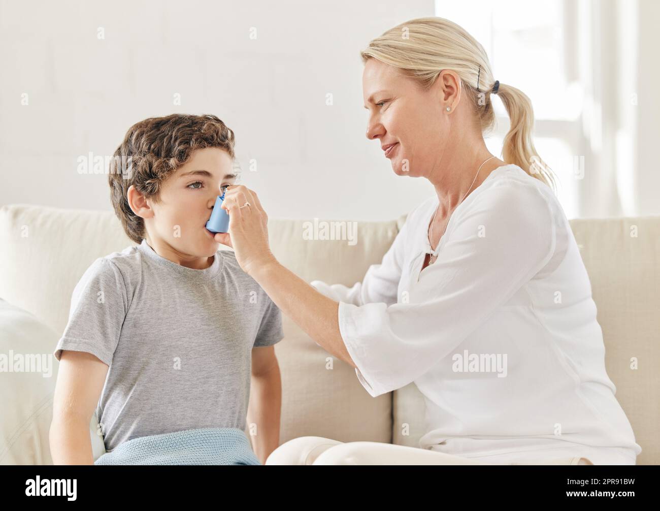 Ein großer Junge mit tiefem Atem. Eine Mutter hilft ihrem Sohn, seinen Inhalator zu Hause auf dem Sofa zu verwenden. Stockfoto