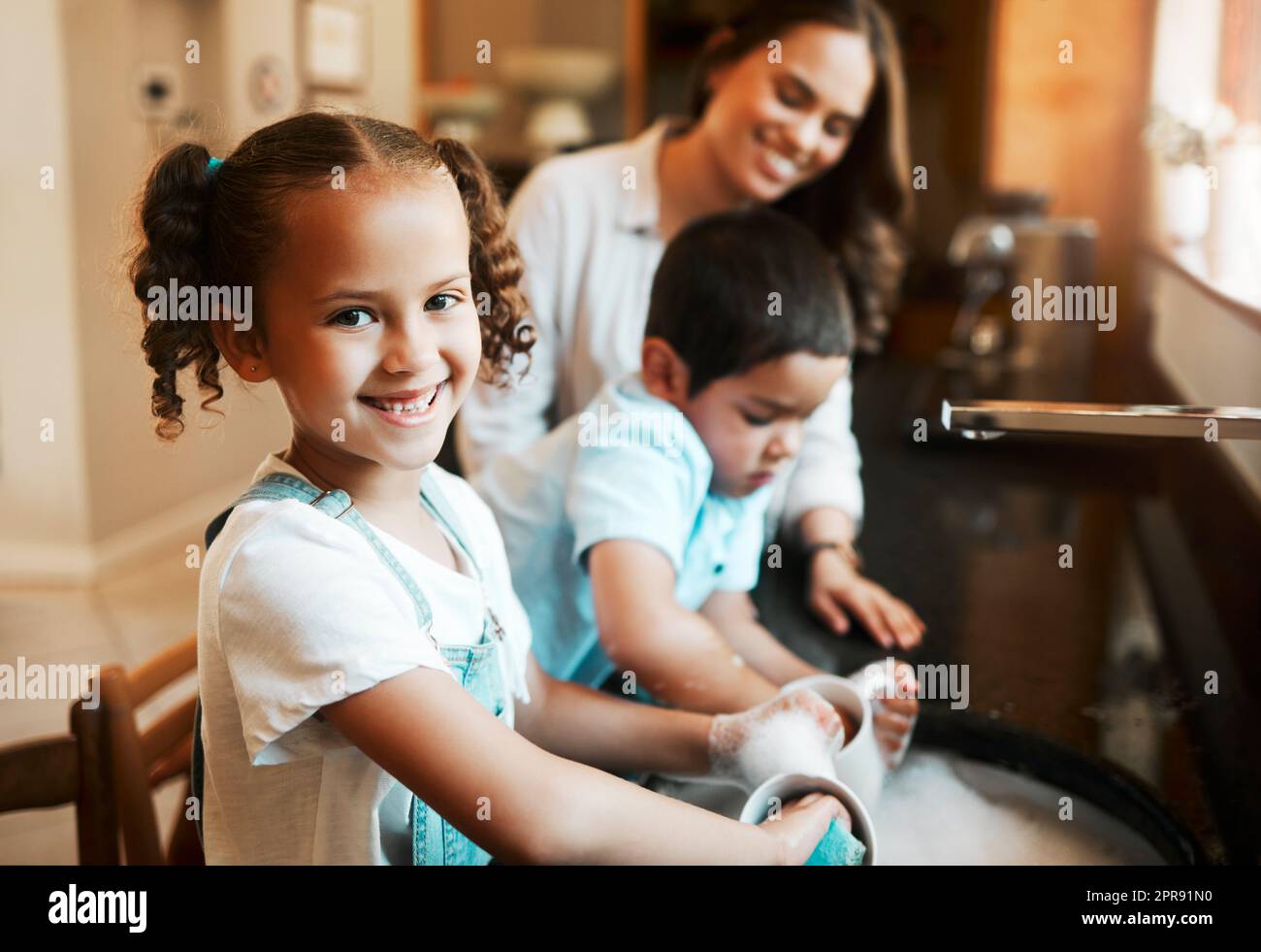 Fröhliches, fröhliches, gemischtes Mädchen, das ihrer Mutter beim Geschirrspülen zuhause in der Küche hilft. Hispanisches Kind lächelt, während es eine Tasse mit Wasser und Seife wäscht. Die Familie hält ihr Haus sauber Stockfoto