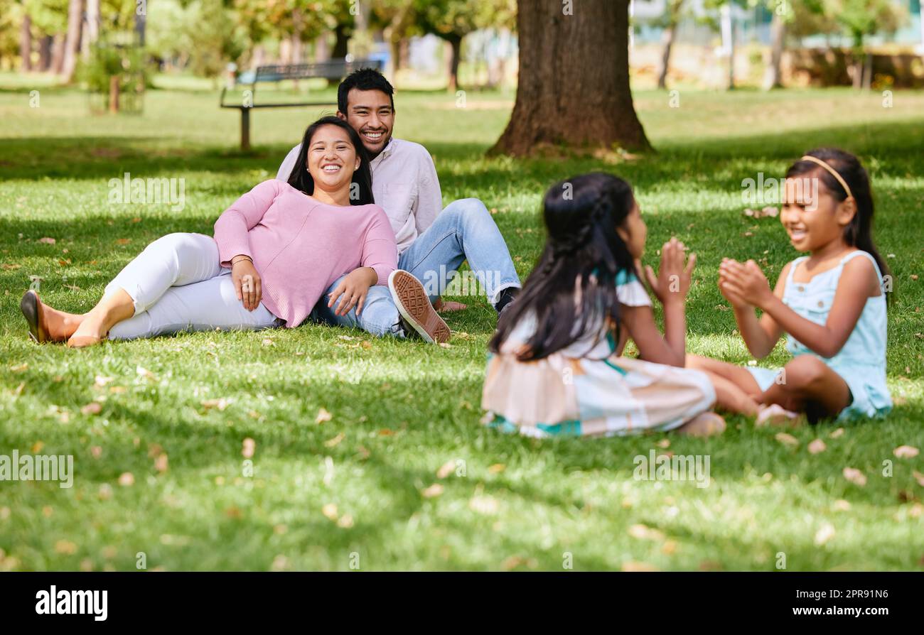 Ein glückliches asiatisches Paar, das draußen auf dem Rasen liegt, liebevolle Eltern genießen eine schöne Zeit mit ihren kleinen Töchtern, die ein Spiel spielen. Ein paar Freundschaften Stockfoto