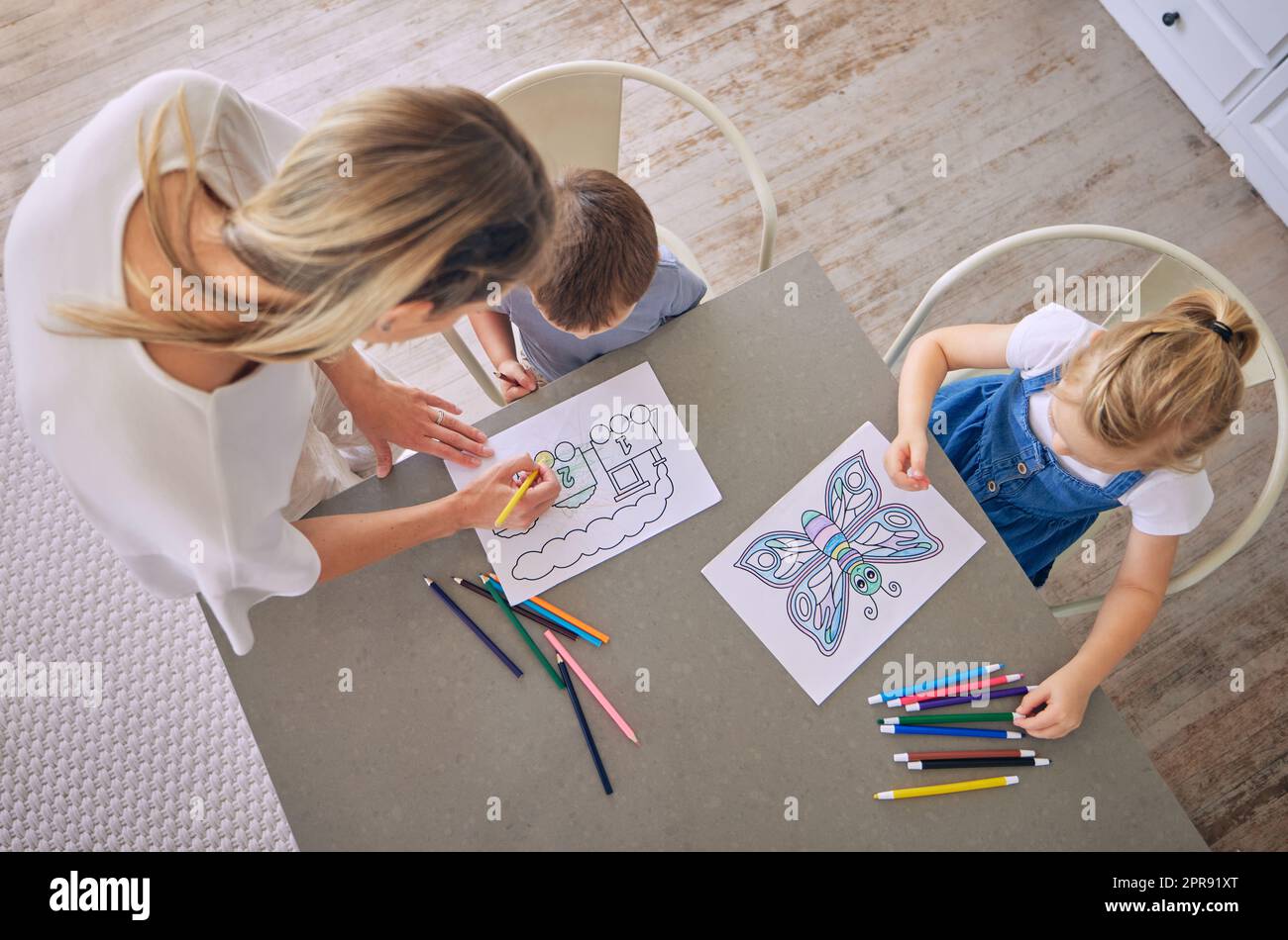 Über dem Bild, wie ein kleines Mädchen und ein Junge mit bunten Bleistiften und Bildern am Tisch sitzen, während sie mit der hilfe der mutter malen. Kaukasische Mutter mit zwei Kindern, die pädagogischen Zeitvertreib genießen und kreativ sind Stockfoto
