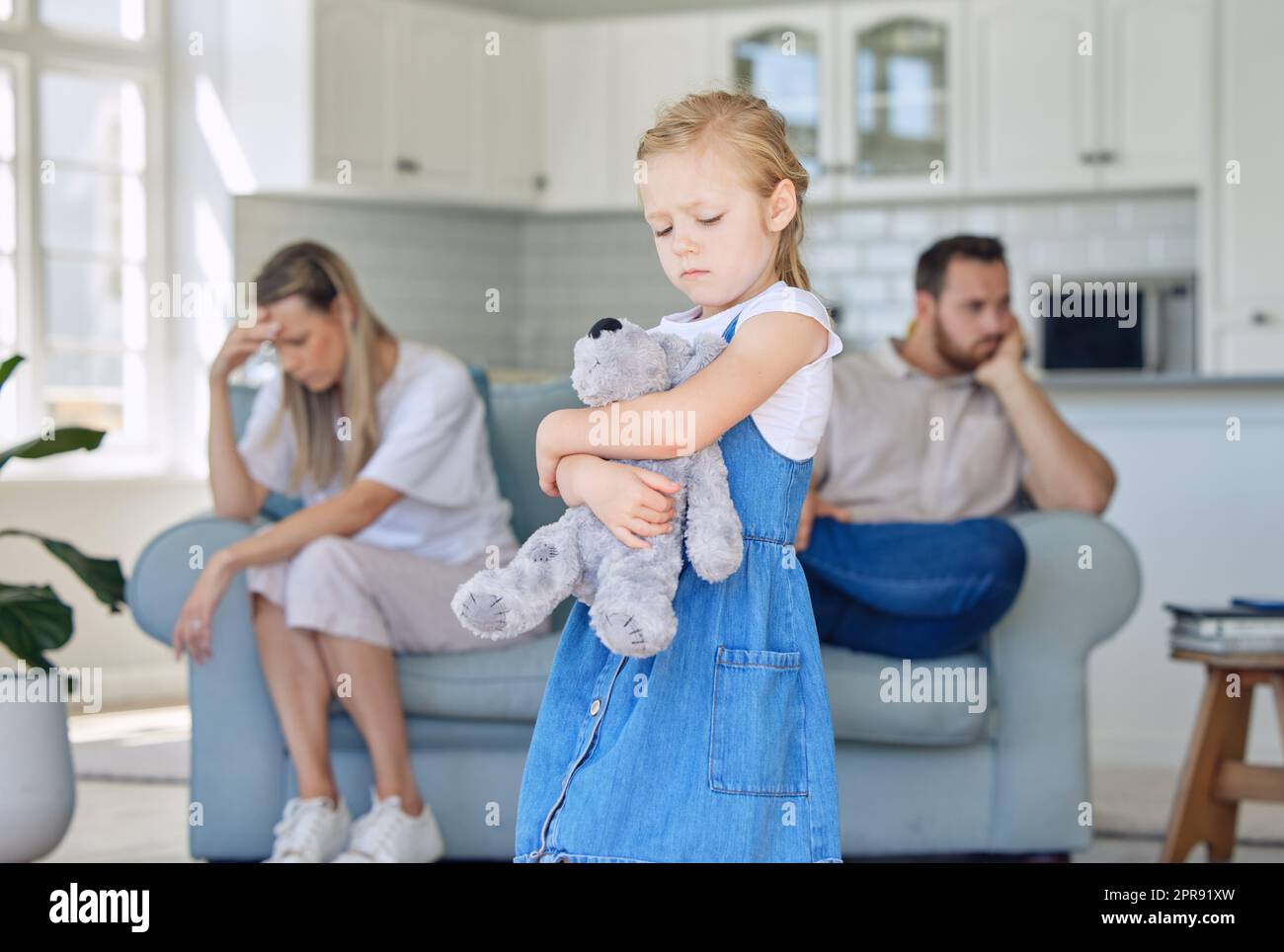 Ein aufgewühltes kleines Mädchen, das ihren Teddybär drückt, während es traurig und deprimiert aussieht, während ihre Eltern im Hintergrund streiten. Dass ihre Eltern sich trennen oder sich scheiden lassen, verursacht Stress Stockfoto