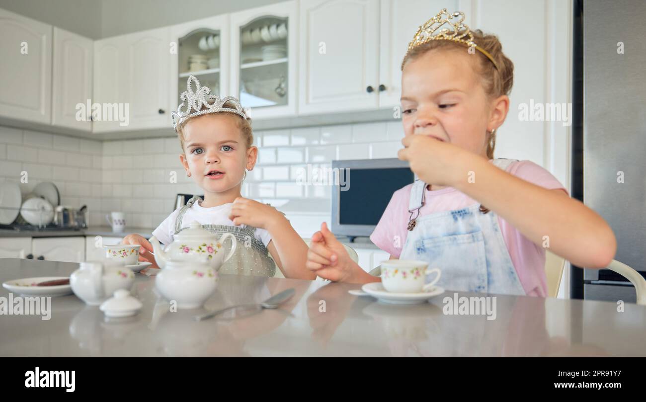 Zwei kleine Mädchen feiern zu Hause eine Teeparty. Geschwisterschwestern tragen Diademe, während sie mit dem Teeset spielen und Kekse am Küchentisch essen. Schwestern verstehen sich gut und spielen zusammen Stockfoto