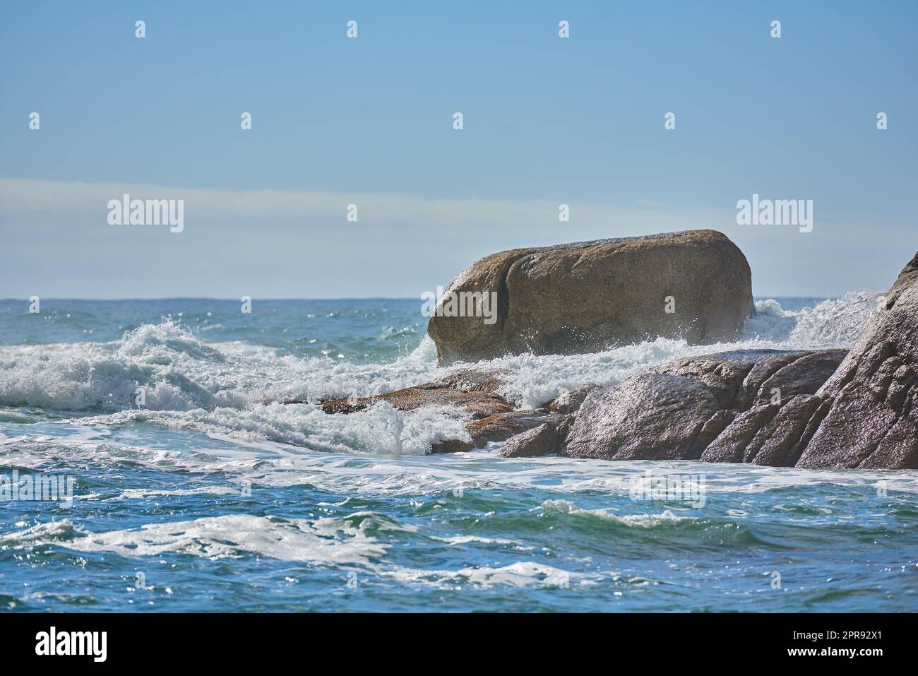 Kopieren Sie den Raum eines turbulenten Meeres mit rauen Gezeiten und zerklüfteten Wellen von starken Winden, die auf große Felsbrocken am Strand mit einem klaren blauen Himmelshintergrund prallen. Felsenküste in Camps Bay, Südafrika Stockfoto