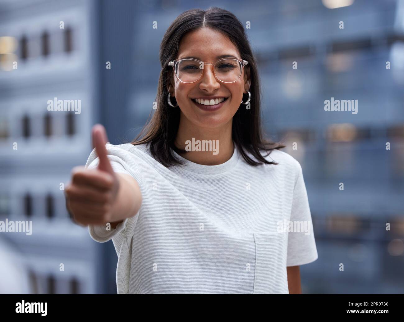 Das Geld wird Ihnen am Ende folgen. Eine junge Geschäftsfrau zeigt Daumen nach oben, während sie vor dem Büro steht. Stockfoto