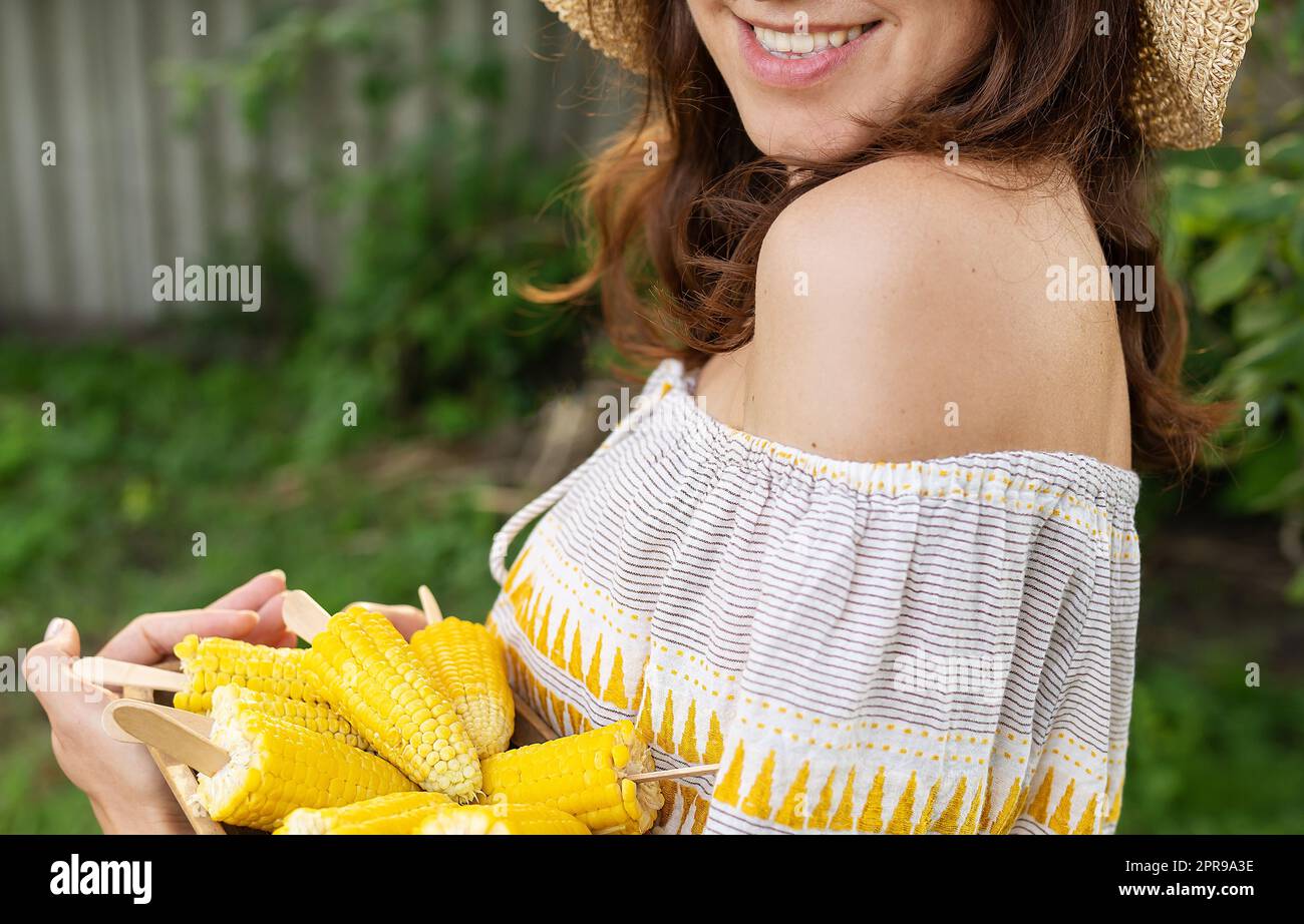 Das Mädchen lächelt in einem Strohhut und hält einen Teller mit gekochtem Mais in der Hand. Das Konzept der Outdoor-Erholung, Barbecue. Stockfoto