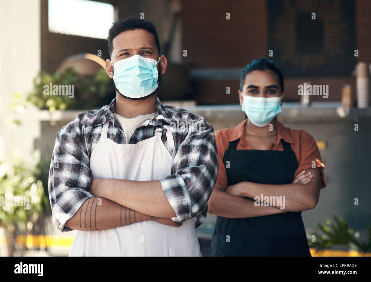 Ihre Gesundheit steht an erster Stelle. Zwei junge Restaurantbesitzer stehen draußen mit gefalteten Armen, während sie Gesichtsmasken tragen. Stockfoto