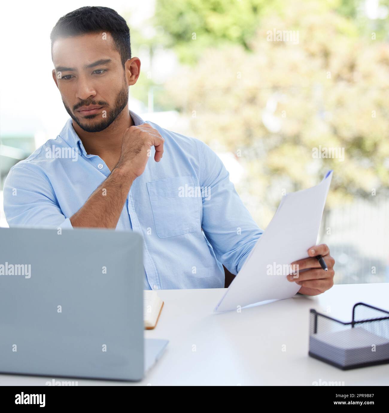 Er übersieht nie die Details. Ein junger Geschäftsmann, der Papierkram durchläuft, während er in einem Büro an einem Laptop arbeitet. Stockfoto