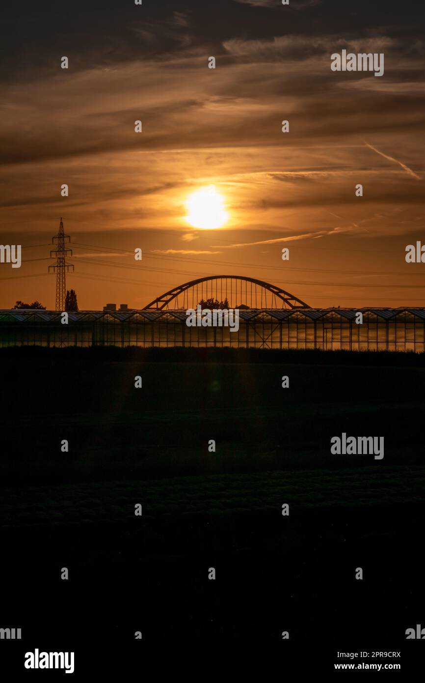 Goldener Sonnenuntergang über Gewächshaussilhouetten mit Brücke und Stromturm für Solarenergie in landwirtschaftlichen Betrieben auf idyllischen Landschaften und ländlicher Landschaft zeigt Glas Gewächshäuser gesundes Gemüse Stockfoto