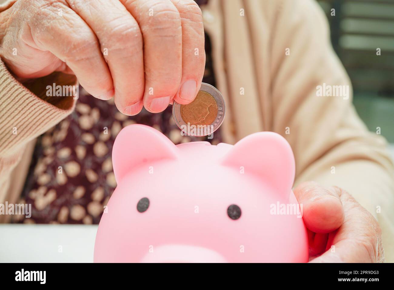 Ältere Frau im Ruhestand, die Münzen auf Sparschwein legt und sich um die monatlichen Ausgaben und die Bezahlung der Behandlungsgebühr sorgt. Stockfoto