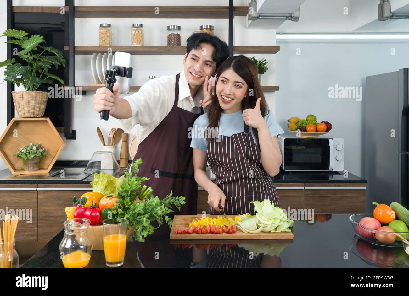 Asiatisches Paar verbringt Zeit zusammen in der Küche. Junge Frau in Schürze kocht Salatgericht, während sein Freund ein Video für einen sozialen Blogger aufnimmt. Moderner Lebensstil, Beziehung und Aktivität. Stockfoto