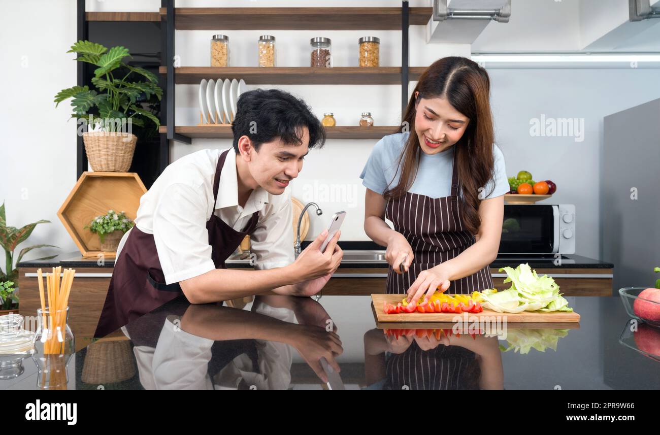 Asiatisches Paar verbringt Zeit zusammen in der Küche. Junge Frau in Schürze kocht Salatgericht, während sein Freund ein Video für einen sozialen Blogger aufnimmt. Moderner Lebensstil, Beziehung und Aktivität Stockfoto