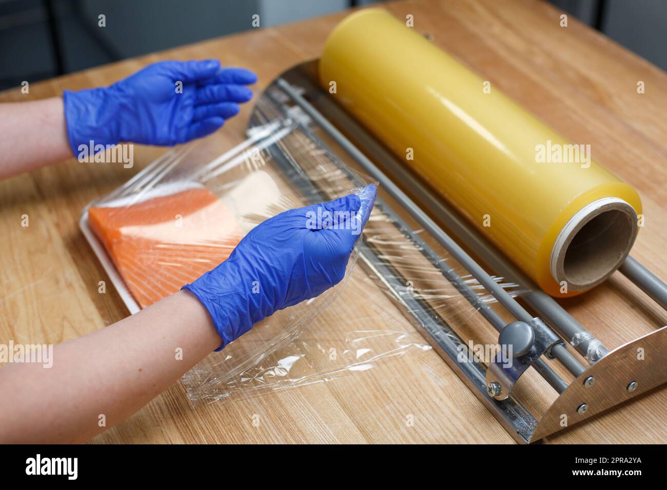 Arbeiter in Einweghandschuhen umwickelt ein Stück Lachsfilet, das auf einer Kunststoffschale in durchsichtiger Folie liegt. Stockfoto