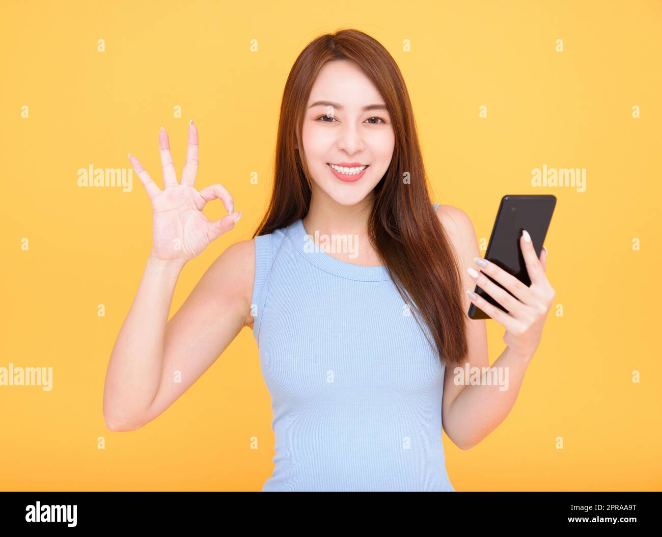 Lächelnde asiatische Frau mit Handy auf gelbem Hintergrund und mit OK-Schild Stockfoto