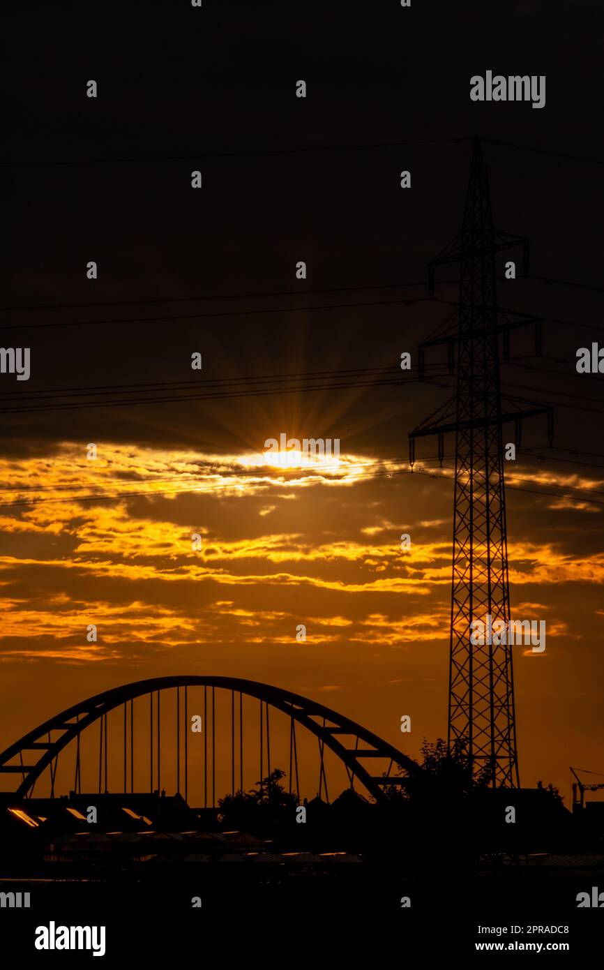 Der goldene Himmel mit Sonnenstrahlen und Linsenerscheinungen zeigt Solarenergie mit der Pylonsilhouette des Stromturms in goldenem Sonnenuntergang und orangefarbenem Himmel für nachhaltige Energie oder erneuerbare Ressourcen von der Dämmerung bis zur Dämmerung Stockfoto