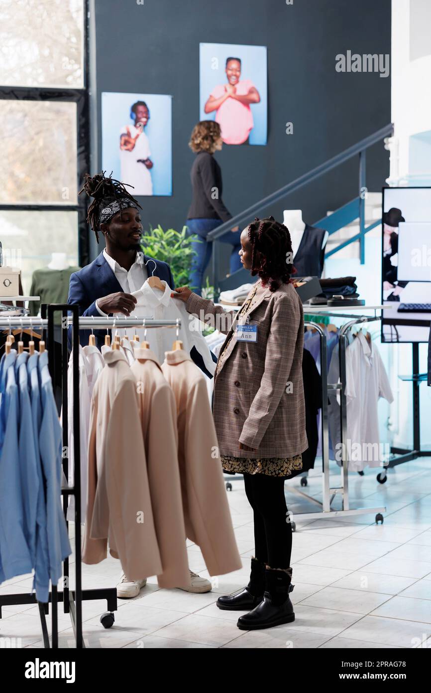 Boutique-Mitarbeiter hilft stilvollen Kunden bei der Auswahl des weißen Hemds während der kommerziellen Aktivitäten. Ein afroamerikanischer Kunde kauft modische Kleidung und Accessoires im Einkaufszentrum. Modekonzept Stockfoto