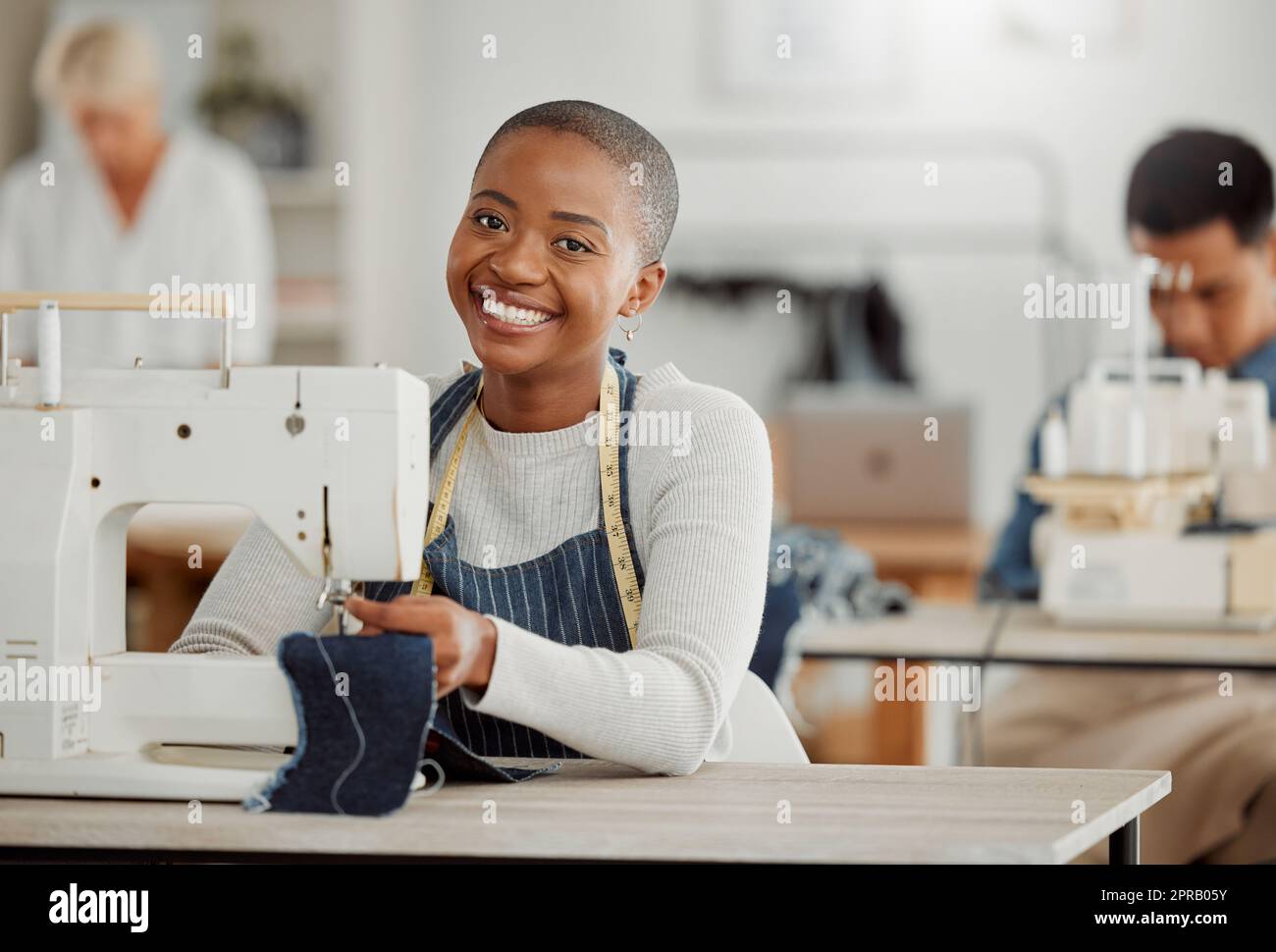 Glücklicher, kreativer schwarzer Modeschüler oder Designer, der im Unterricht an einer Nähmaschine sitzt und lächelt, die an Kleidung arbeitet. Isoliertes Porträt eines wunderschönen afroamerikanischen Schneiders in einer Fabrik oder einem Studio. Stockfoto