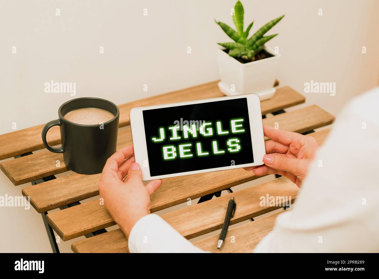 Handschriftlich auf dem Schild "Jingle Bells". Internetkonzept weltweit berühmtester traditioneller Weihnachtssong Frau mit Tablet und aktuellen Daten auf dem Tisch mit Kaffee, Stift und Pflanzen Stockfoto