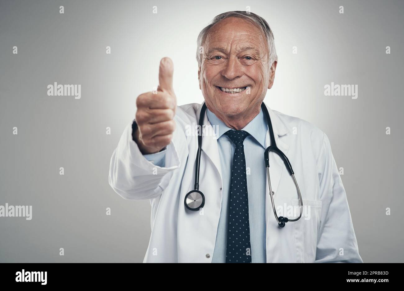 Jahre später liebe ich immer noch meinen Job. Ein älterer, männlicher Arzt in einem Studio, der vor grauem Hintergrund Daumen nach oben gibt. Stockfoto