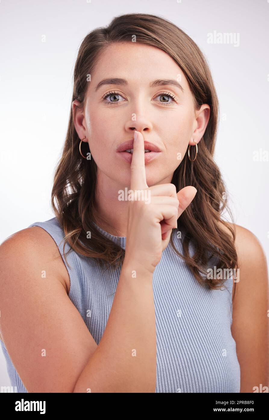 Ihr sagt es niemandem besser. Studioportrait einer jungen Frau mit dem Finger auf den Lippen vor grauem Hintergrund. Stockfoto