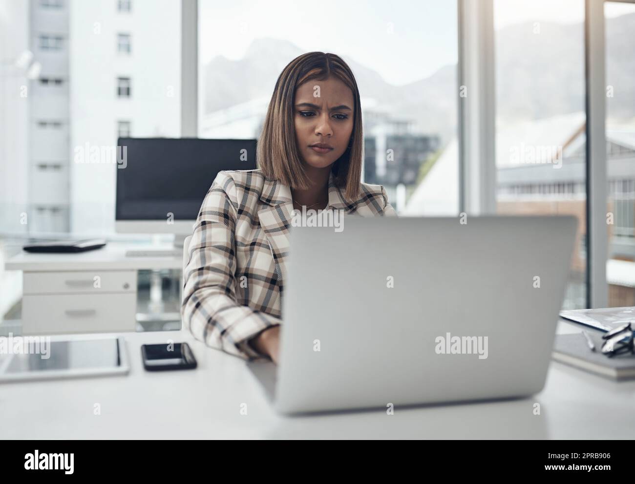 Etwas scheint nicht ganz richtig zu sein: Eine attraktive junge Geschäftsfrau, die allein in ihrem Büro sitzt und verwirrt aussieht, während sie ihren Laptop benutzt. Stockfoto