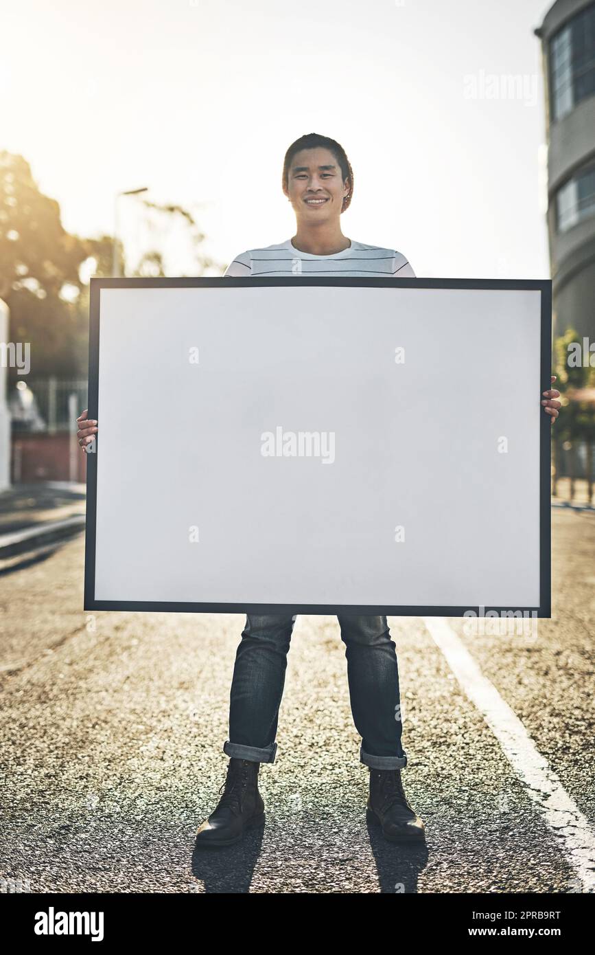 Junger Mann mit leerem Schild, Brett oder Plakat, während er draußen auf der Straße der Stadt steht. Porträt eines glücklichen, lächelnden und fröhlichen Mannes, der eine Anzeige zeigt oder befördert Stockfoto