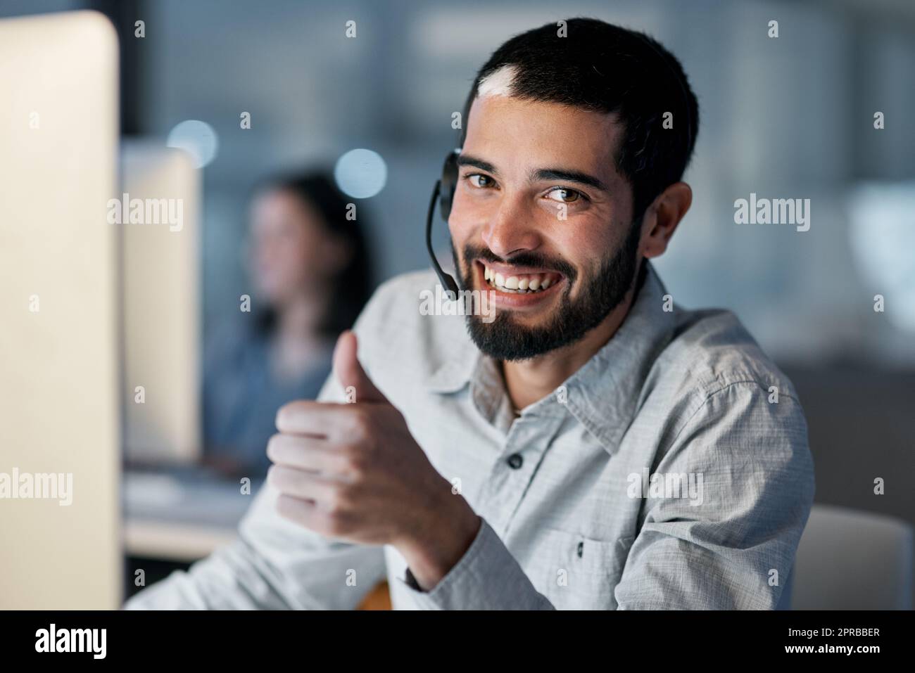 Ein junger Mann, der in einem modernen Büro spät in der Nacht ein Headset und einen Computer benutzt, zeigt den Daumen nach oben. Stockfoto