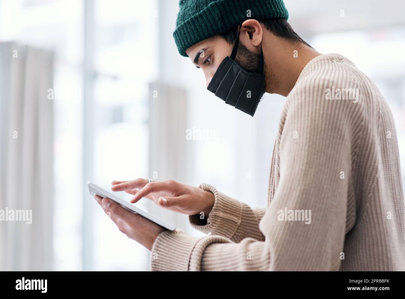 Die Arbeit muss weitergehen. Ein maskierter junger Mann, der ein digitales Tablet benutzt, während er von zu Hause aus arbeitet. Stockfoto