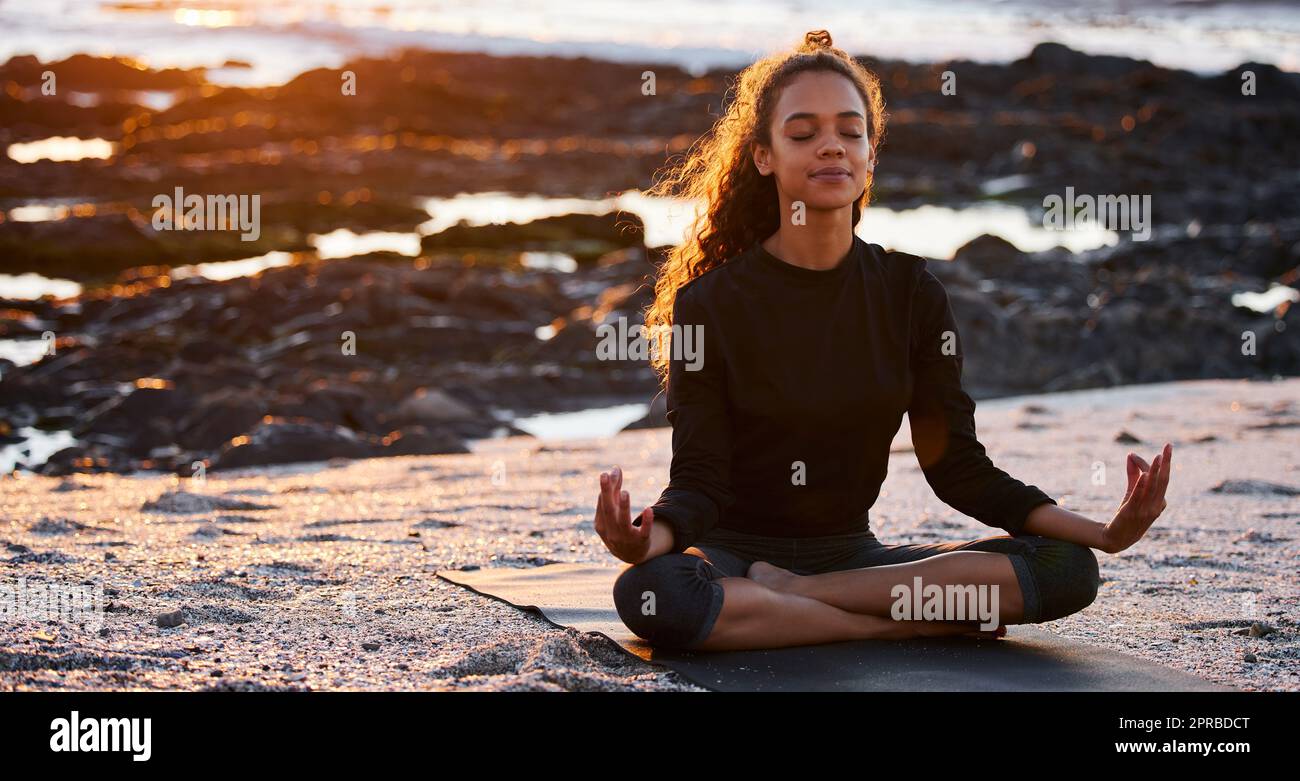 Dankbarkeit bringt positive Veränderungen. Eine attraktive junge Frau, die allein auf einer Matte sitzt und bei Sonnenuntergang am Strand meditiert. Stockfoto