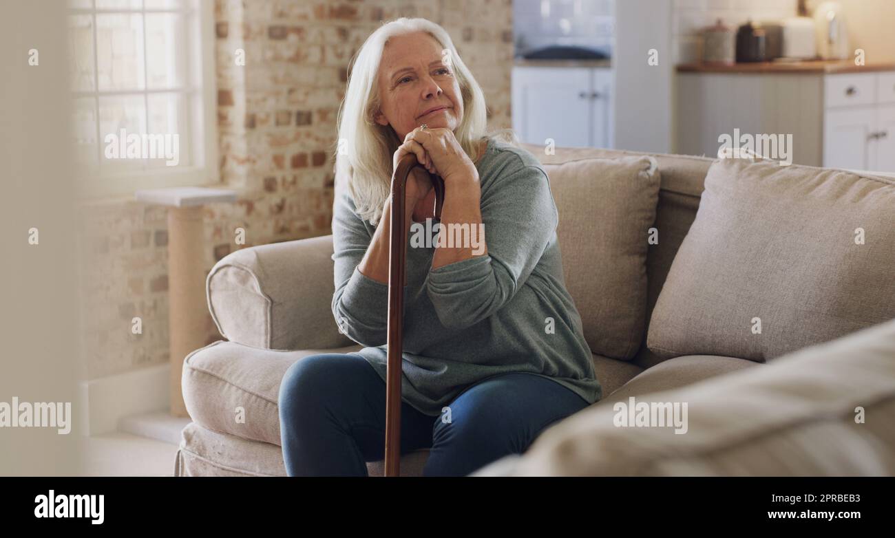 Ich habe die ganze Zeit in der Welt zum Nachdenken. Eine ältere Frau, die allein auf dem Sofa zu Hause sitzt und besinnlich aussieht, während sie ihren Spazierstock hält. Stockfoto