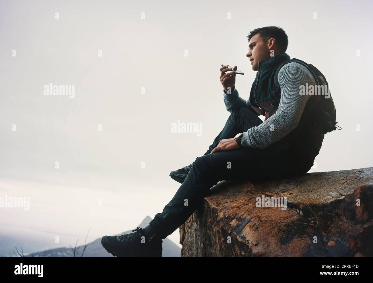 Ein junger Mann, der auf einem Handy spricht, während er auf einer Bergklippe sitzt. Stockfoto