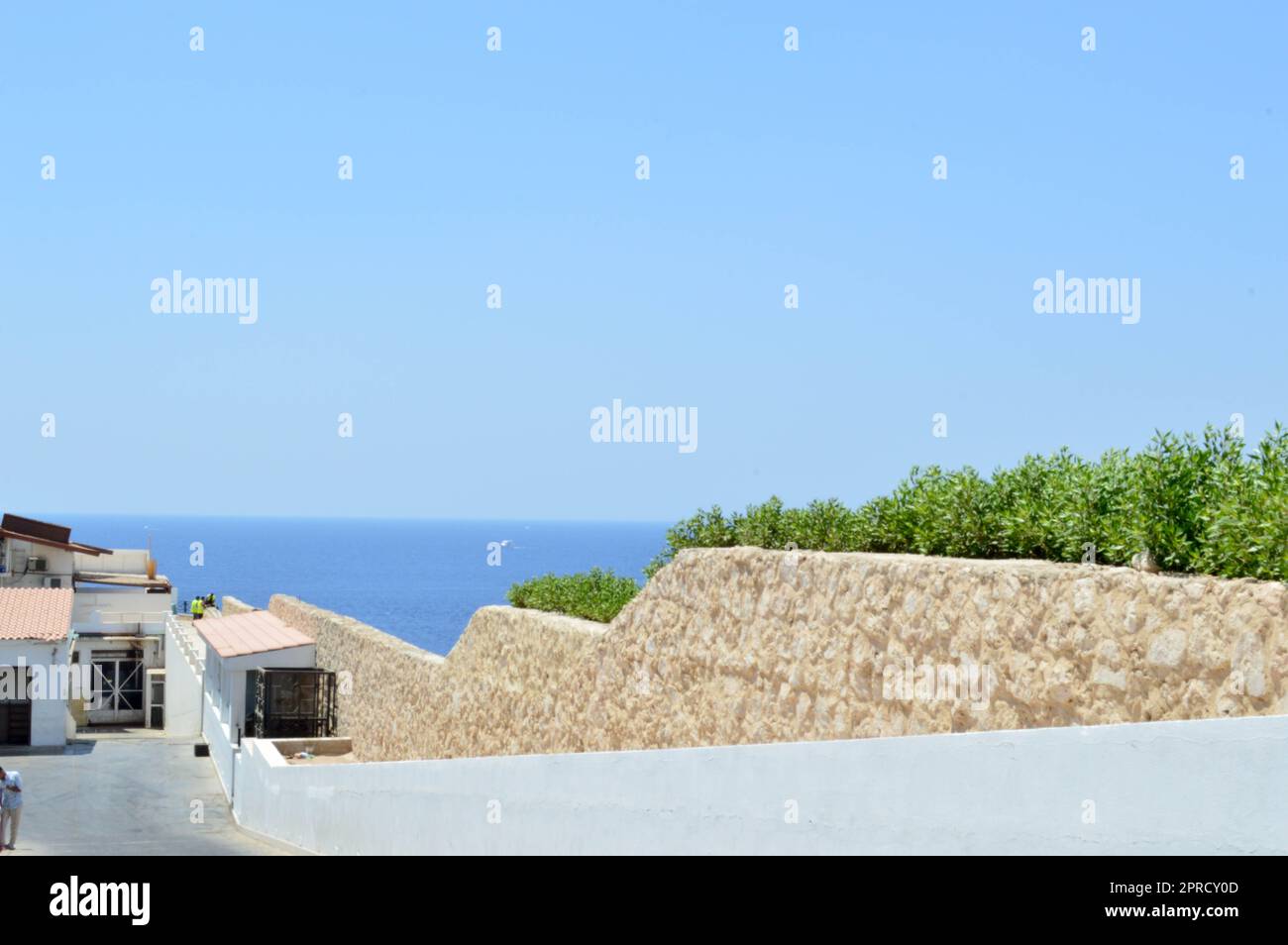 Eine wunderschöne Steinmauer hinunter zum Meer mit blauem Wasser in einem tropischen Resort und ein weißes Gebäude mit einem Dach aus roten Schindeln und grünem Busch Stockfoto